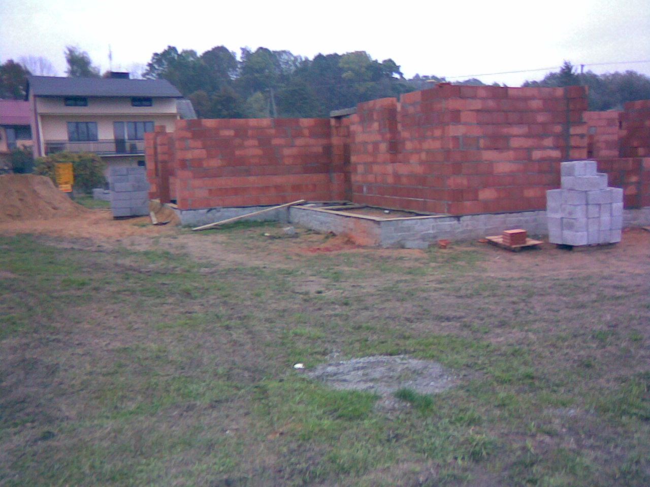 Wznoszenie murów z czerwonej cegły na fundamencie betonowym, widok na budowę domu jednorodzinnego w trakcie realizacji, sterta bloczków obok.